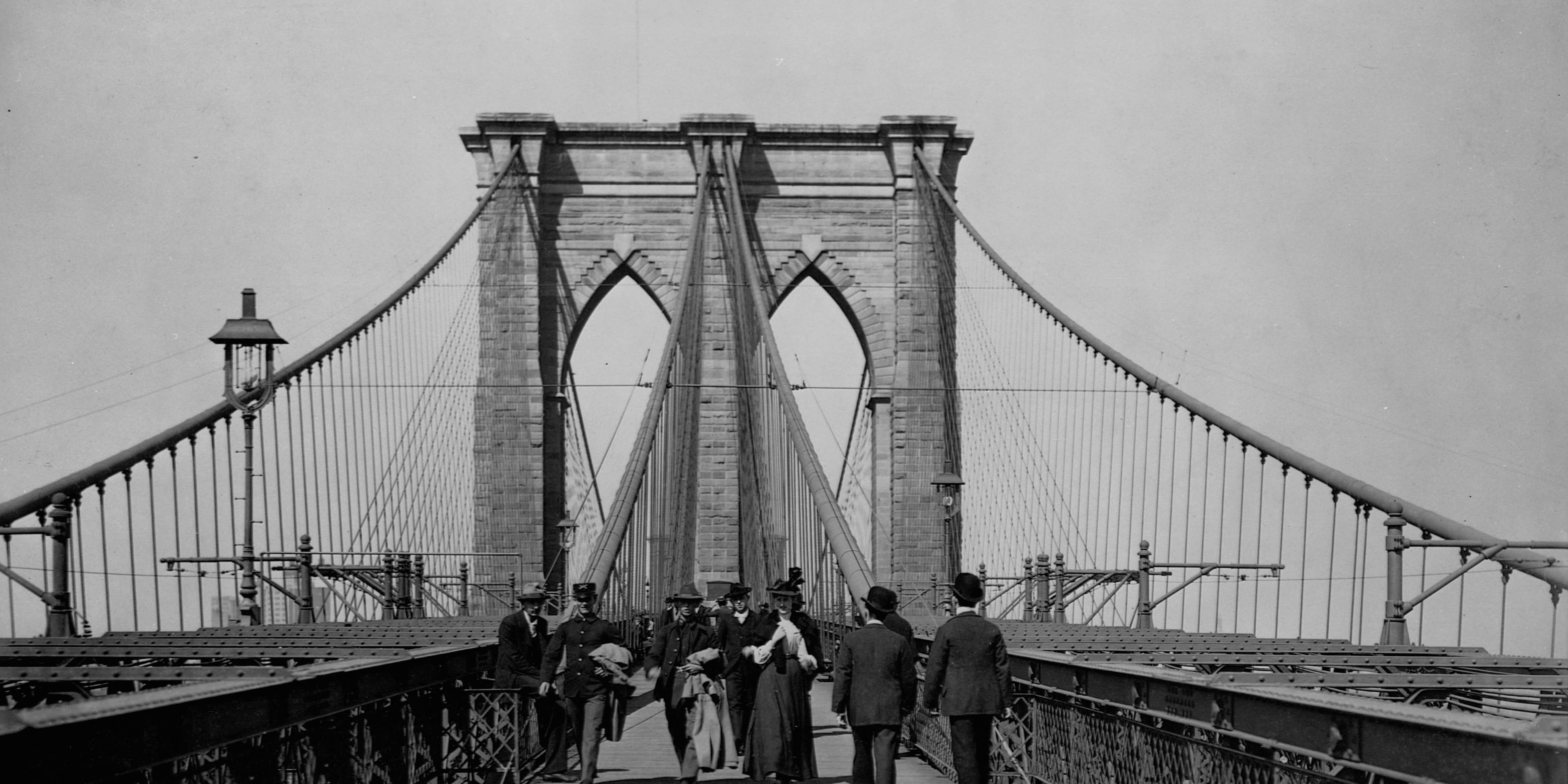 Men and women stroll along the promenade deck of the Brooklyn Bridge.