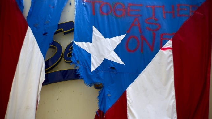 A damaged Puerto Rican national flag spray painted hangs from the facade of a business in San Juan after Hurricane Maria. Puerto Rico has so far been concentrated largely in San Juan, and many outside the capital say they've received little or no help. (Credit: Ramon Espinosa/AP Photo)
