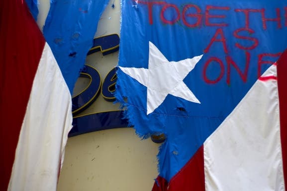 A damaged Puerto Rican national flag spray painted hangs from the facade of a business in San Juan after Hurricane Maria. Puerto Rico has so far been concentrated largely in San Juan, and many outside the capital say they've received little or no help. (Credit: Ramon Espinosa/AP Photo)