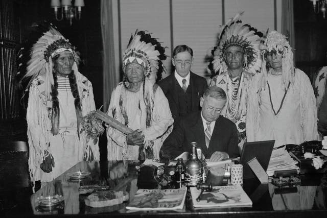 Government official in suit sitting at a desk signing a document, as a group of men in <a href=